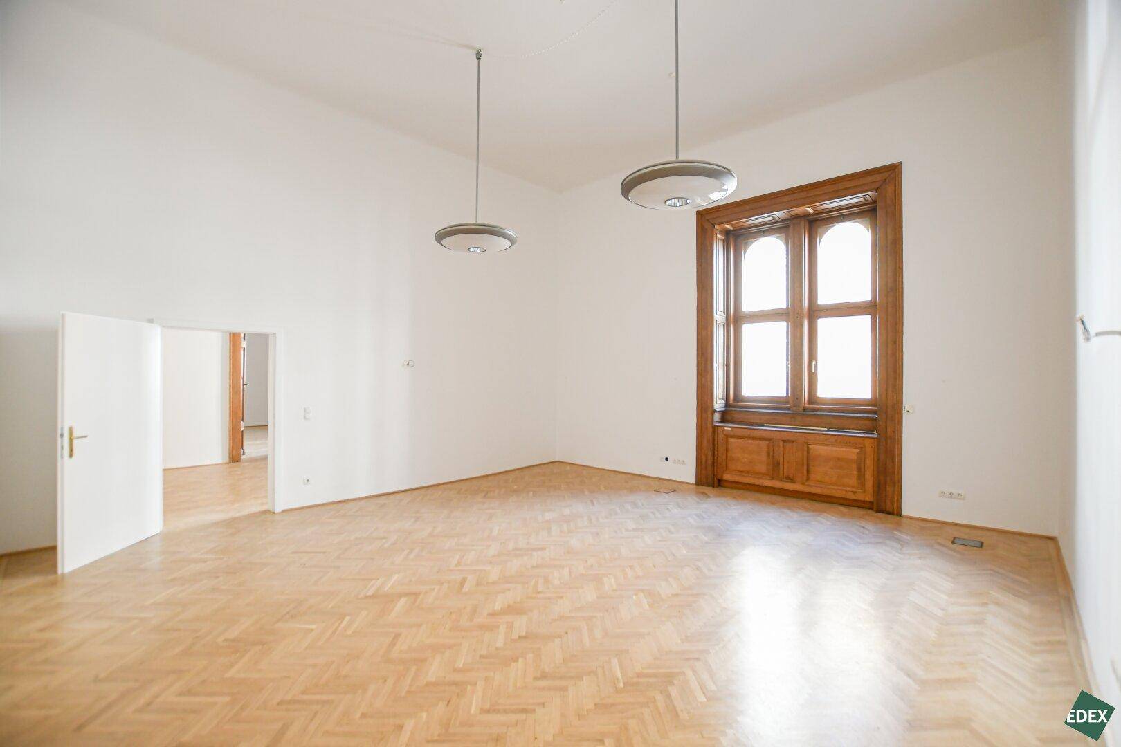 Geräumiger, heller Büroraum mit hohen Decken, elegantem Fischgrätparkett und einem großen Holzfenster.