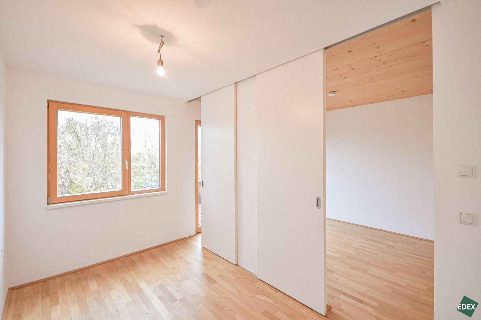 Schlafzimmer mit Holzboden, Holzdecke und einem großen Fenster, das viel Tageslicht hereinlässt.