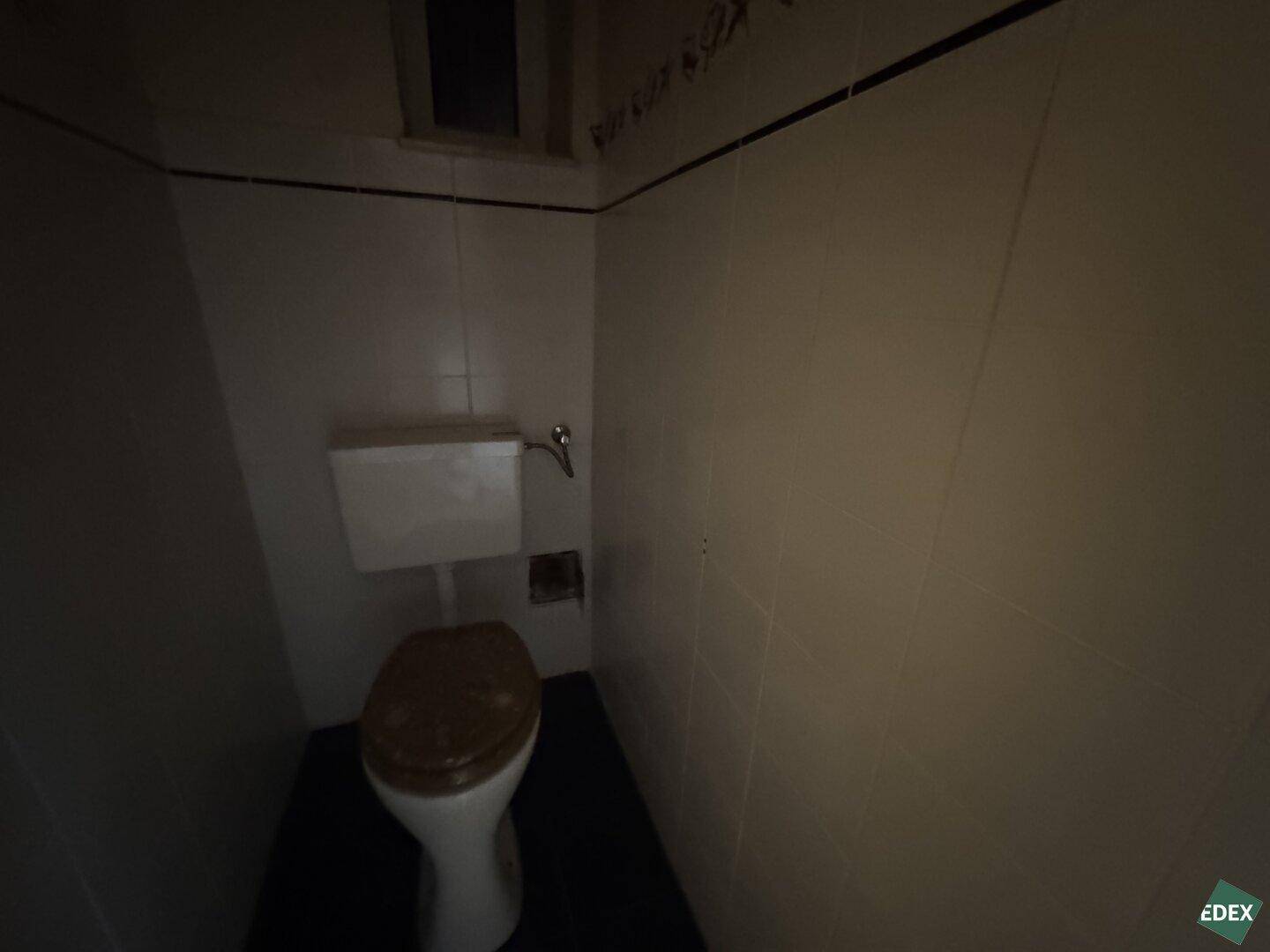 Kleines WC mit Toilette und dunklem Boden, funktional aber renovierungsbedürftig.