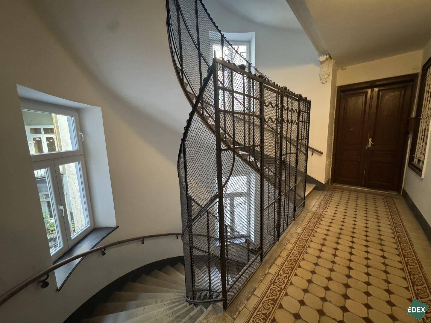 Historisches Treppenhaus mit kunstvollem Metallgeländer und großen Fenstern, Altbaucharme.