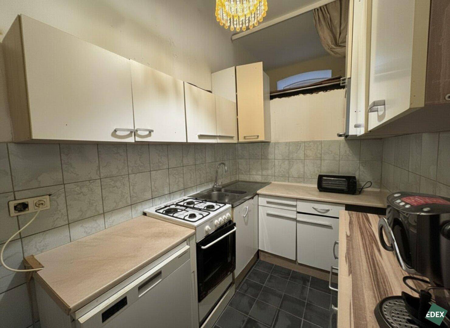 Kleine Küche mit Gasherd, Spüle und weißen Hängeschränken, typisch für eine ältere Wohnung.
