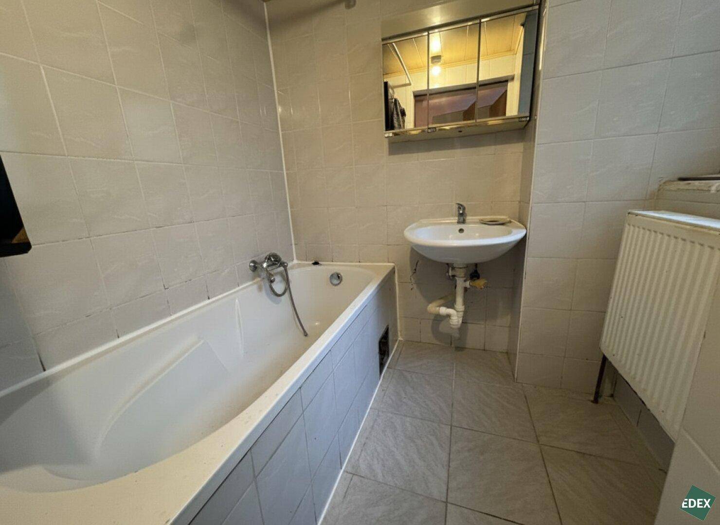 Badezimmer mit Badewanne, Waschbecken und hellen Fliesen, ausgestattet mit einem Heizkörper.