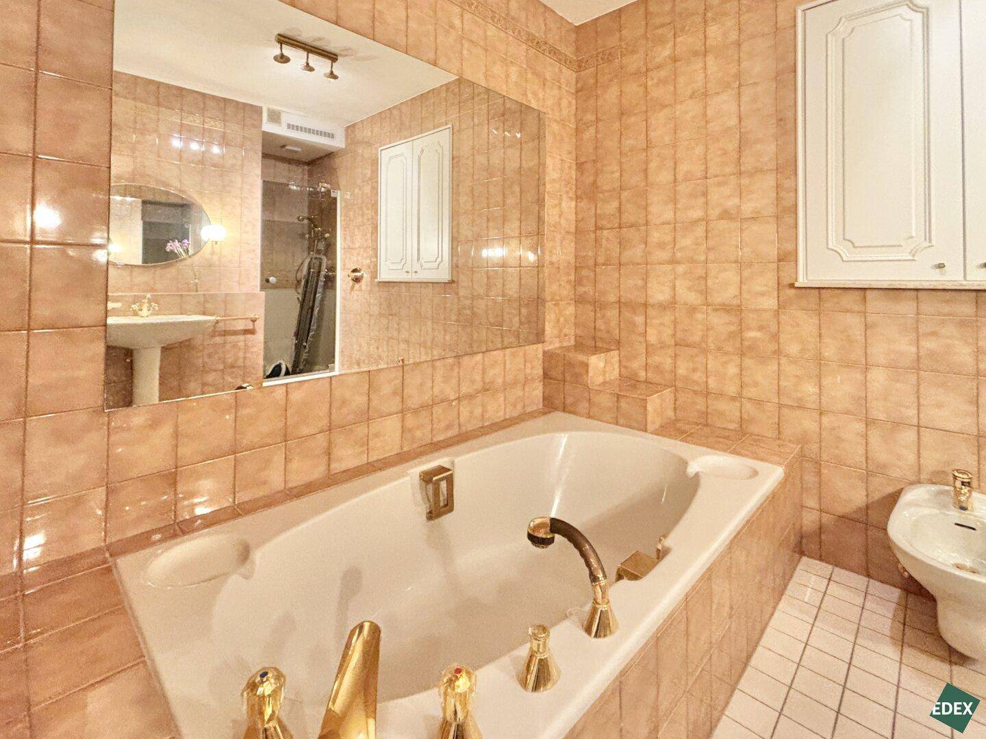 Eingebaute Badewanne mit goldenen Armaturen und großem Wandspiegel im gefliesten Badezimmer.