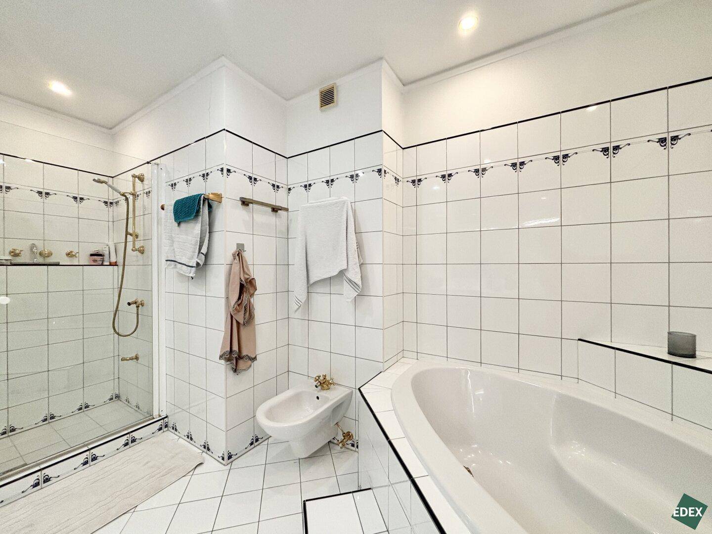 Badezimmer mit Badewanne, Duschkabine, Bidet und weißen Fliesen mit blauen Bordüren im 80er-Jahre-Stil.