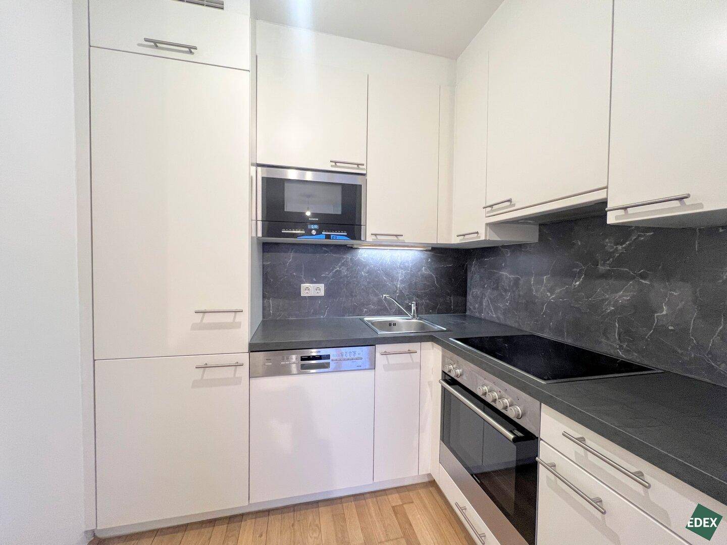 Kompakte, moderne Küchenzeile mit weißen Schränken, dunkler Arbeitsplatte und Einbaugeräten.