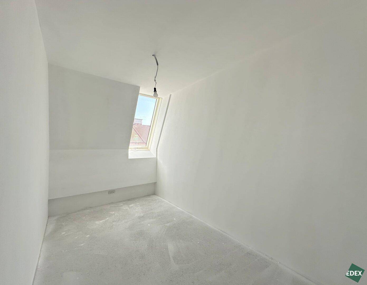 Leeres Zimmer im Dachgeschoss mit einem einzelnen Dachfenster und hellen Wänden.