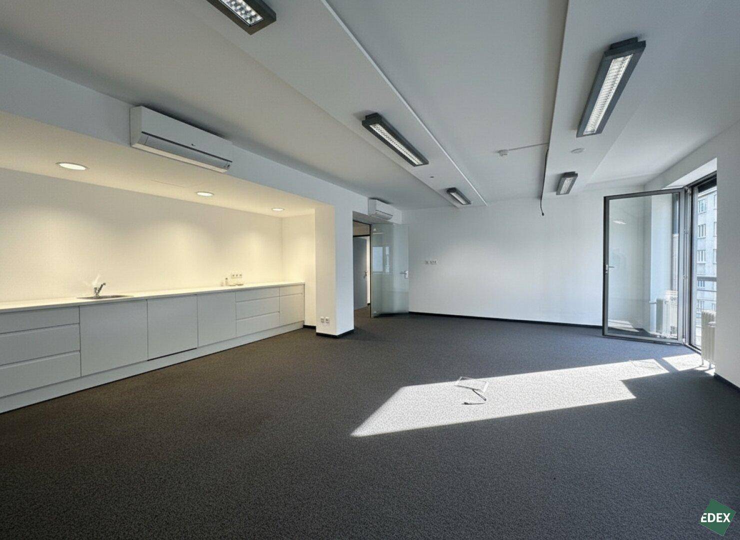 Helles Büro mit Teppichboden, integrierter Küchenzeile und Zugang zu einem Balkon.