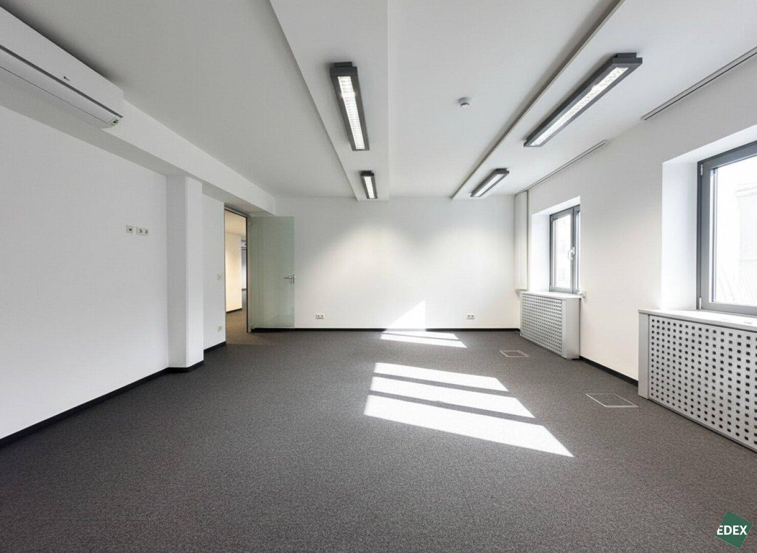 Leeres, helles Büro mit Teppichboden, großen Fenstern und moderner Beleuchtung.