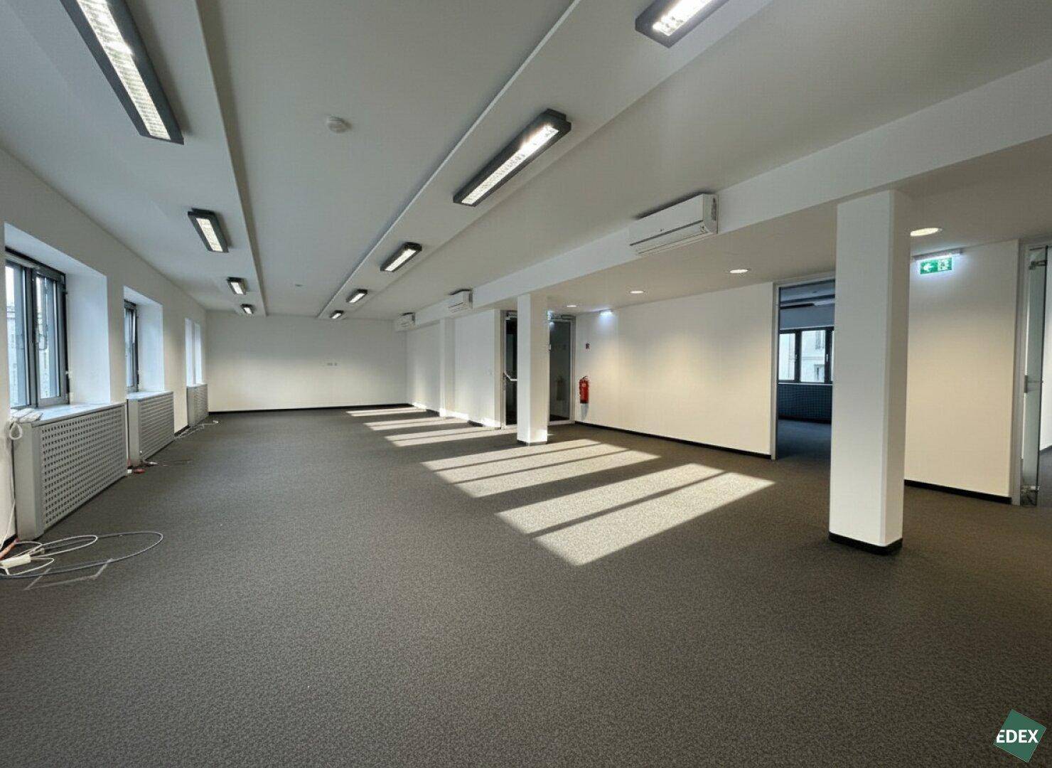 Weitläufiges, leeres Büro mit Teppichboden, Säulen und vielen Fenstern für natürliches Licht.