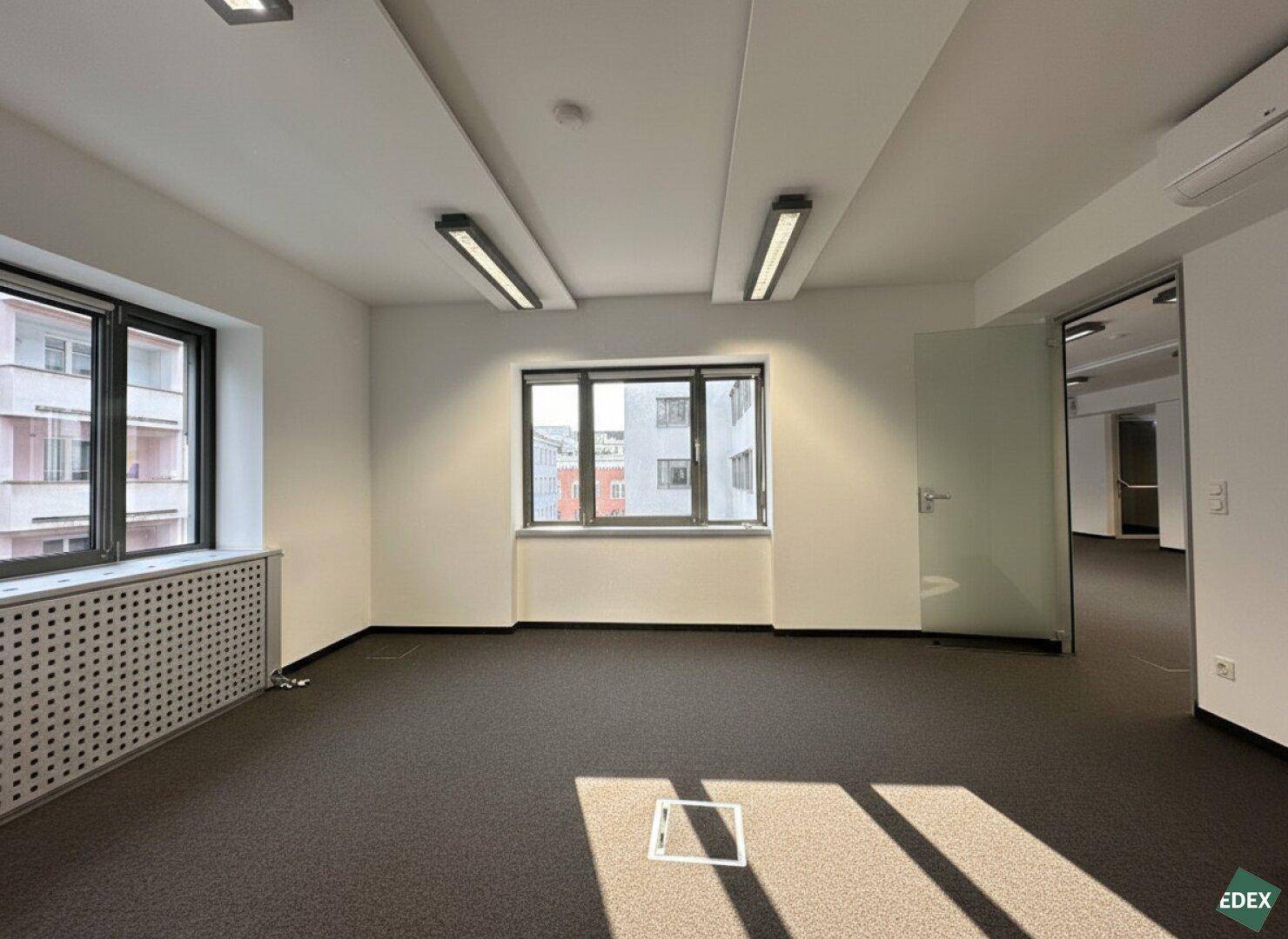 Helles Büro mit Teppichboden, großen Fenstern und Blick auf die umliegenden Gebäude.