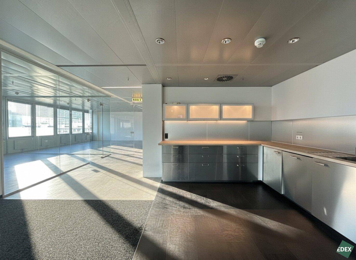 Moderne Teeküche in einem Bürogebäude mit Edelstahloberflächen, Holztheke und Blick in den offenen Bürobereich.