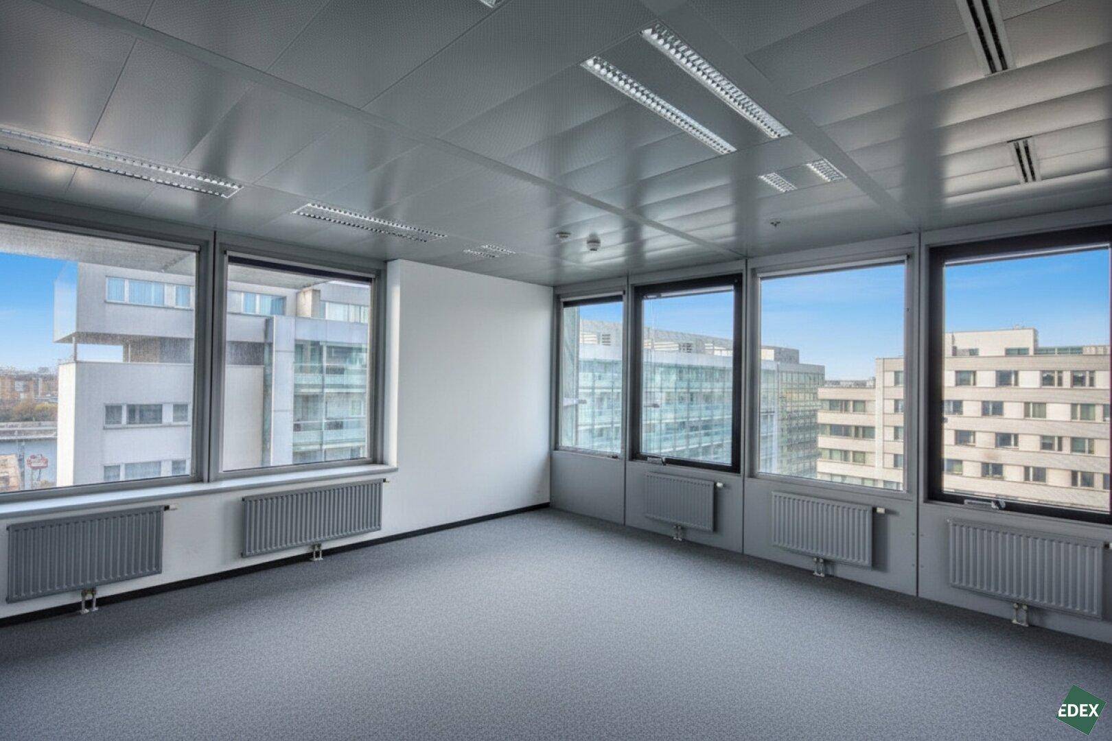 Heller Büroraum mit großen Fenstern, Teppichboden und Blick auf umliegende Gebäude.