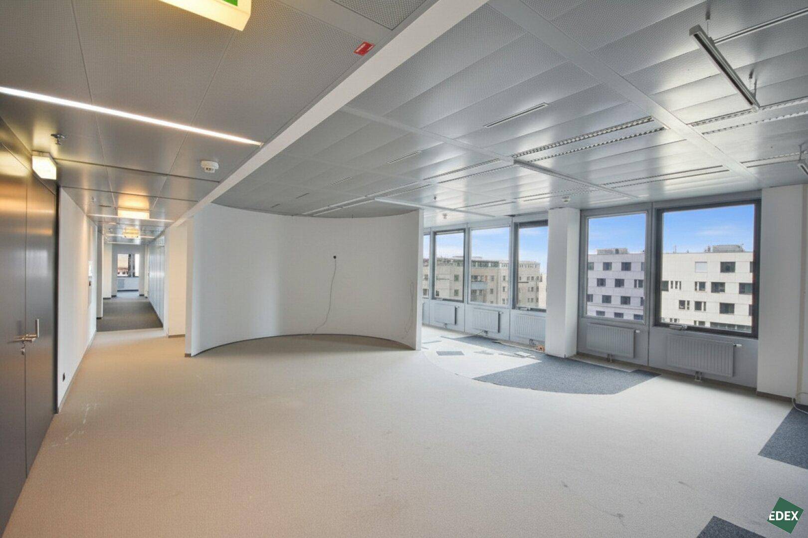 Geräumiger, heller Bürobereich mit großen Fenstern, Teppichboden und Blick auf die Stadt.