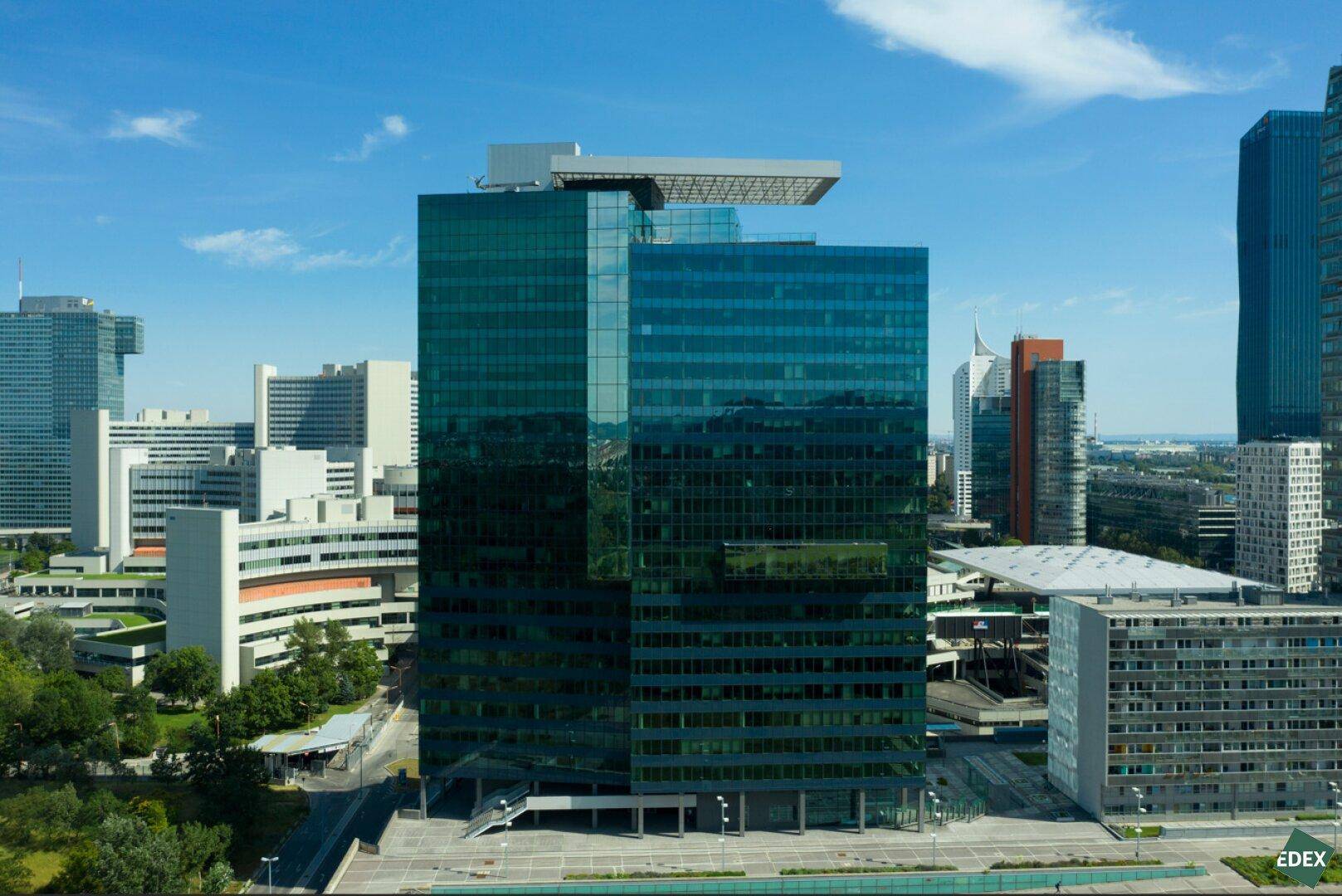 Panoramablick auf ein modernes Hochhaus mit Glasfassade, umgeben von anderen Gebäuden und Grünflächen.