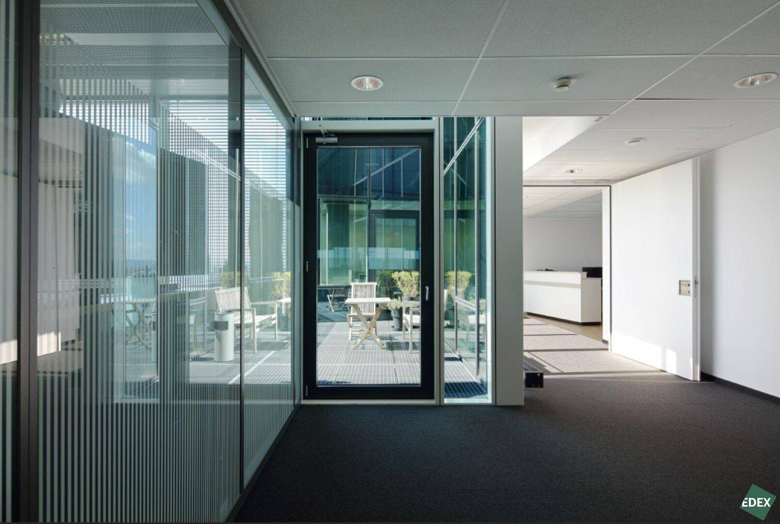 Heller Bürogang mit Glaswänden und Zugang zu einer möblierten Außenterrasse.