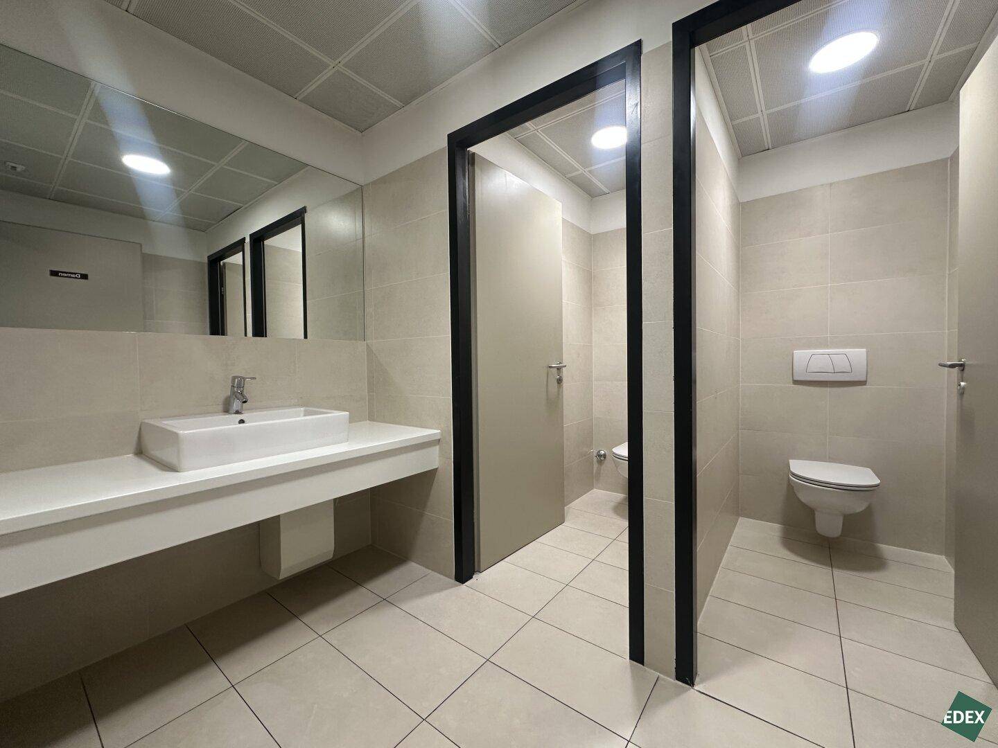 Moderne Toilettenanlage mit mehreren Kabinen, Waschbecken und großem Spiegel, in hellen Fliesen gehalten.