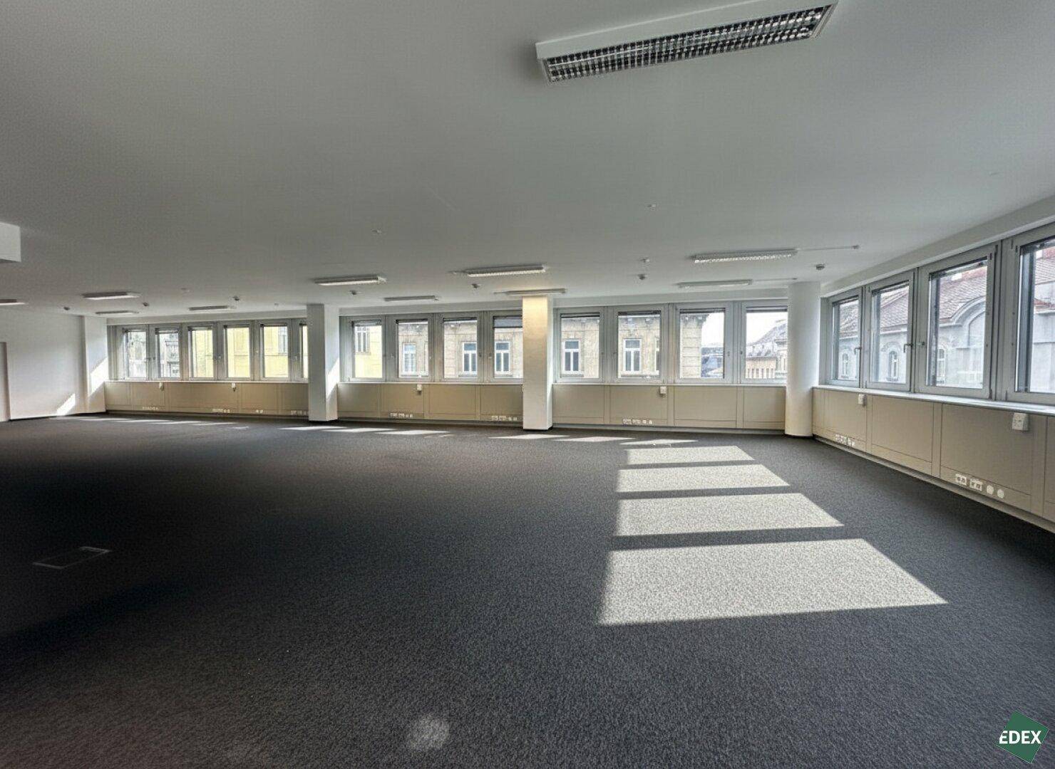 Heller Büroraum mit dunklem Teppichboden und einer langen Fensterfront, die den Raum mit Licht füllt.