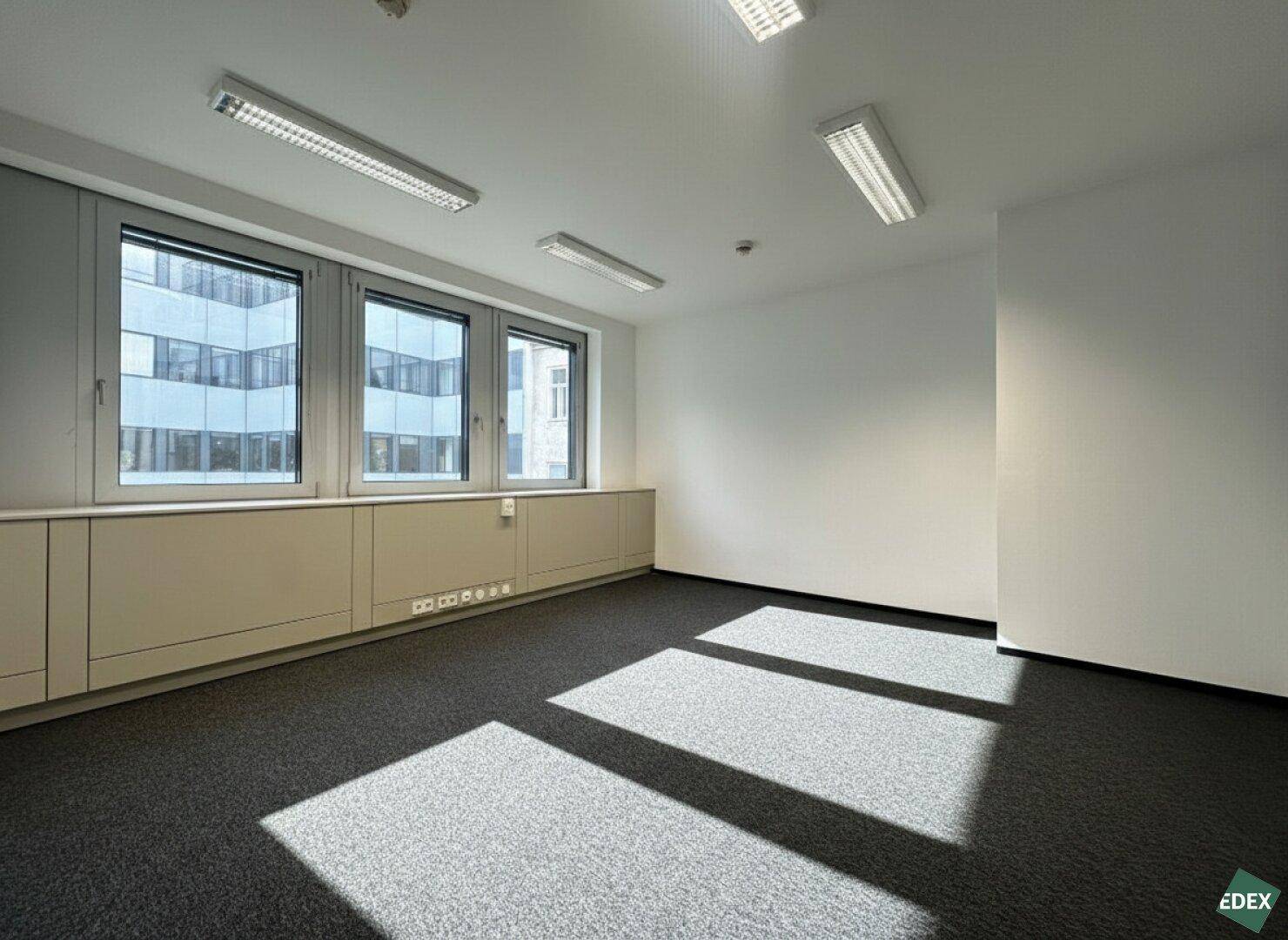 Heller Büroraum mit Teppichboden und einer Fensterfront, die einen Blick auf die umliegenden Gebäude bietet.