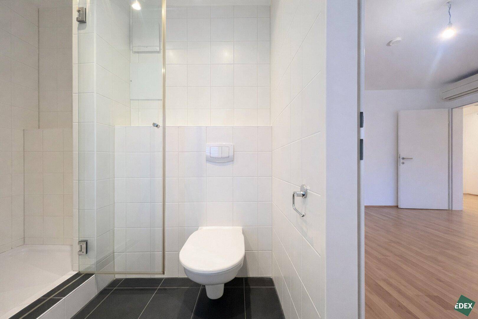 Helles Badezimmer mit wandmontierter Toilette und Duschbereich, direkt vom Wohnraum zugänglich.