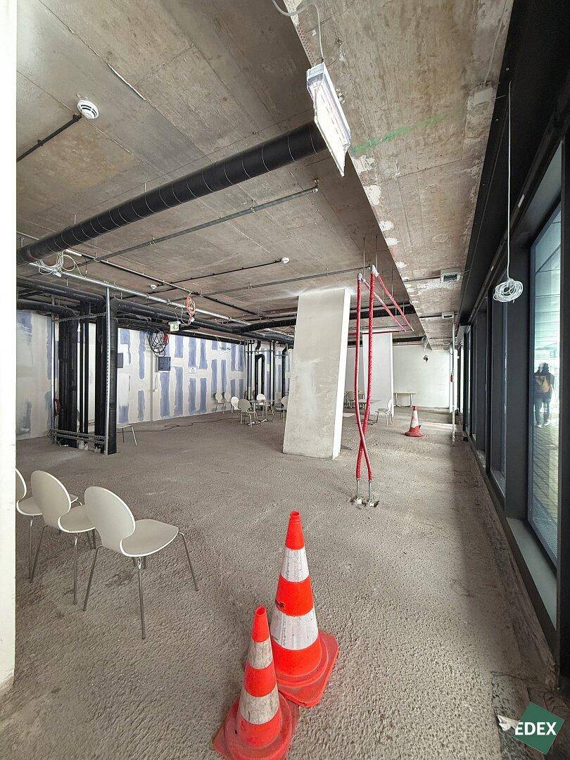 Großer, unfertiger Gewerberaum mit Betonboden, freiliegenden Leitungen und großen Fenstern, bereit für den Innenausbau.