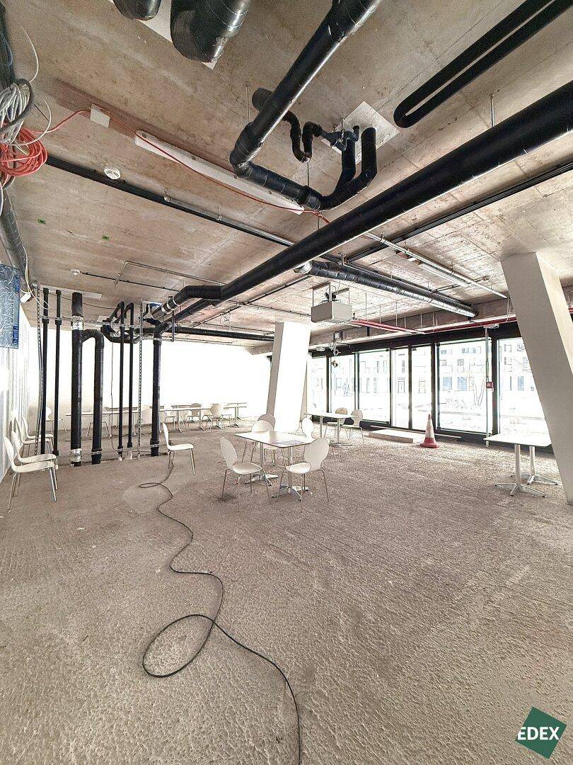 Weitläufiger, unfertiger Gewerberaum mit Betonboden, freiliegenden Leitungen und großen Fenstern, bereit für den Ausbau.