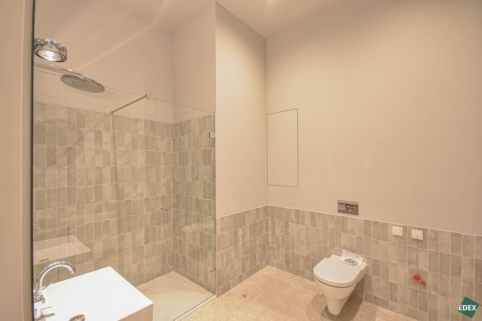 Modernes Badezimmer mit begehbarer Dusche, Wand-WC und hellgrauen Fliesen an Wänden und Boden.