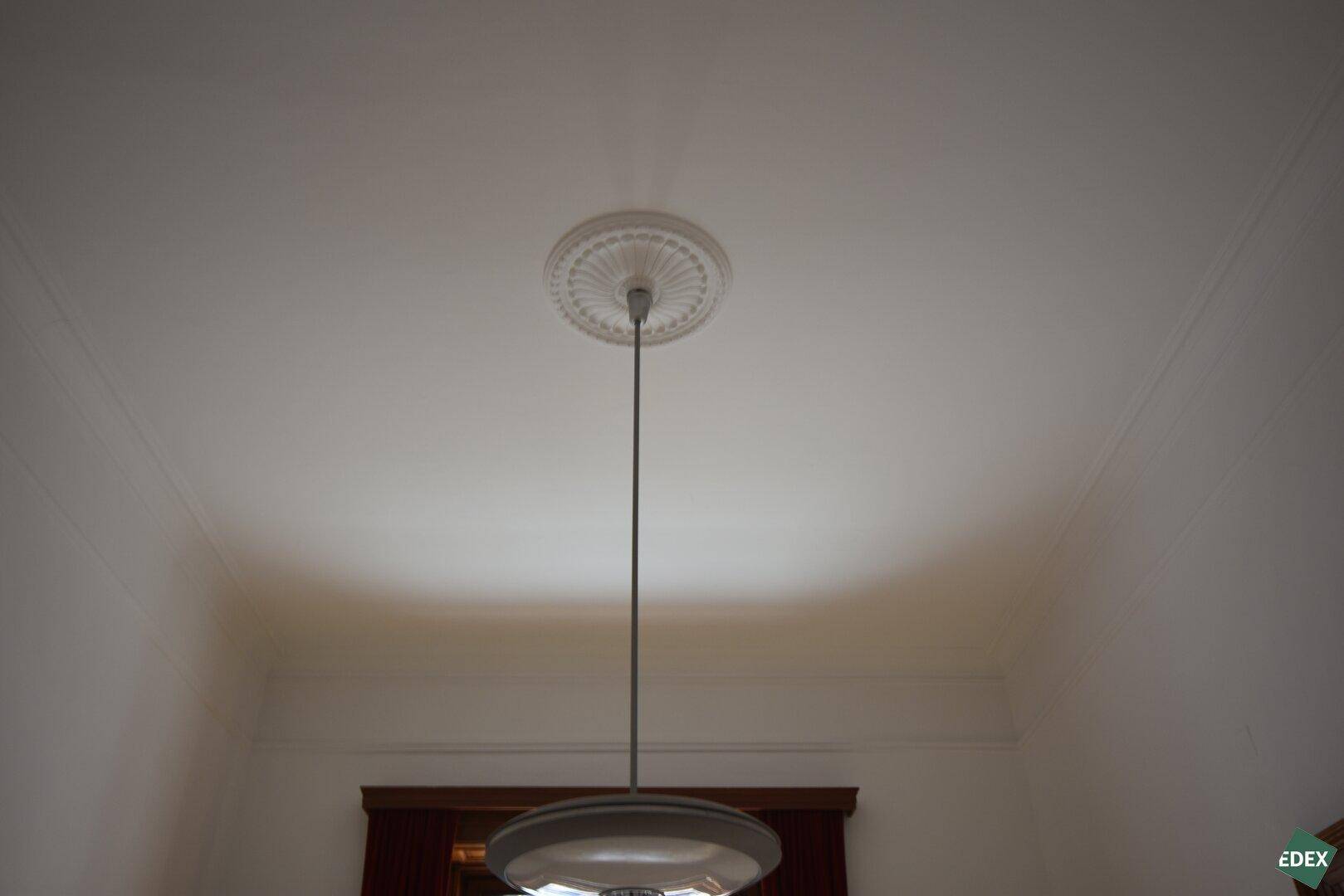 Decke mit klassischem Stuckmedaillon und einer modernen Hängelampe in einem hohen Raum.