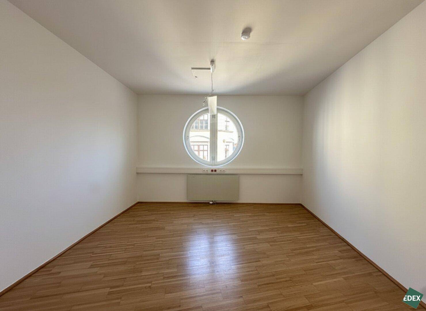 Büroraum mit einzigartigem Rundfenster, Heizkörper und klassischem Parkettboden, bietet besonderen Charme.