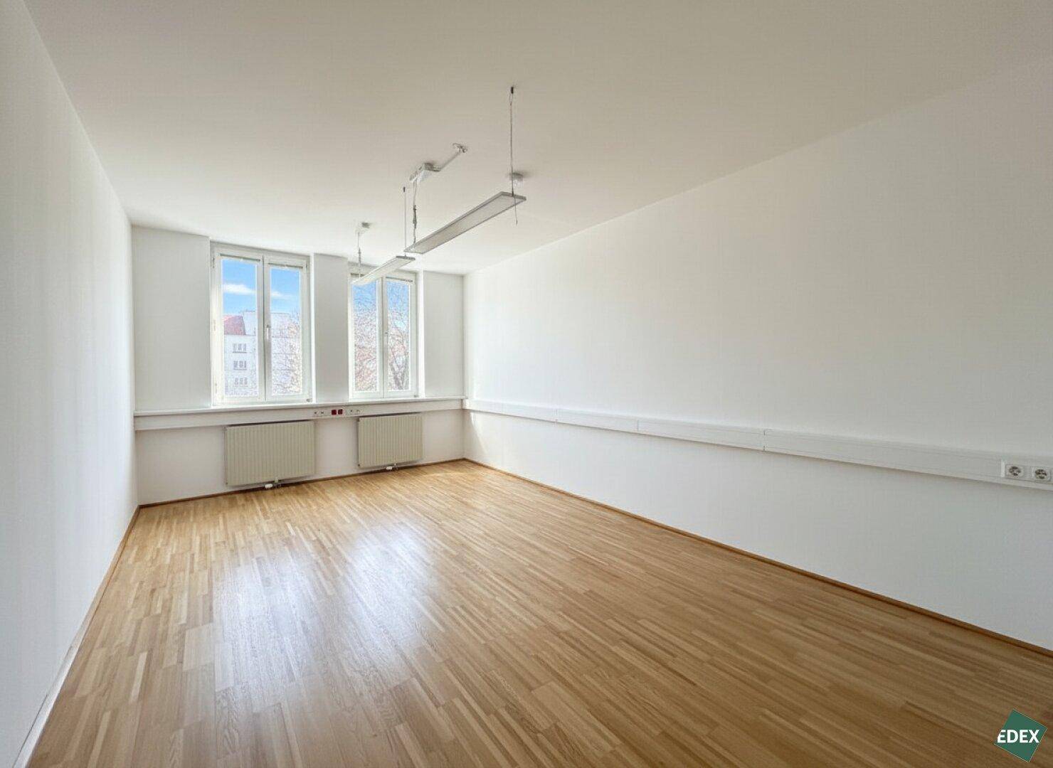 Kleinerer Büroraum mit zwei Fenstern, Heizkörpern und hellem Parkettboden, ideal für Einzelbüros.