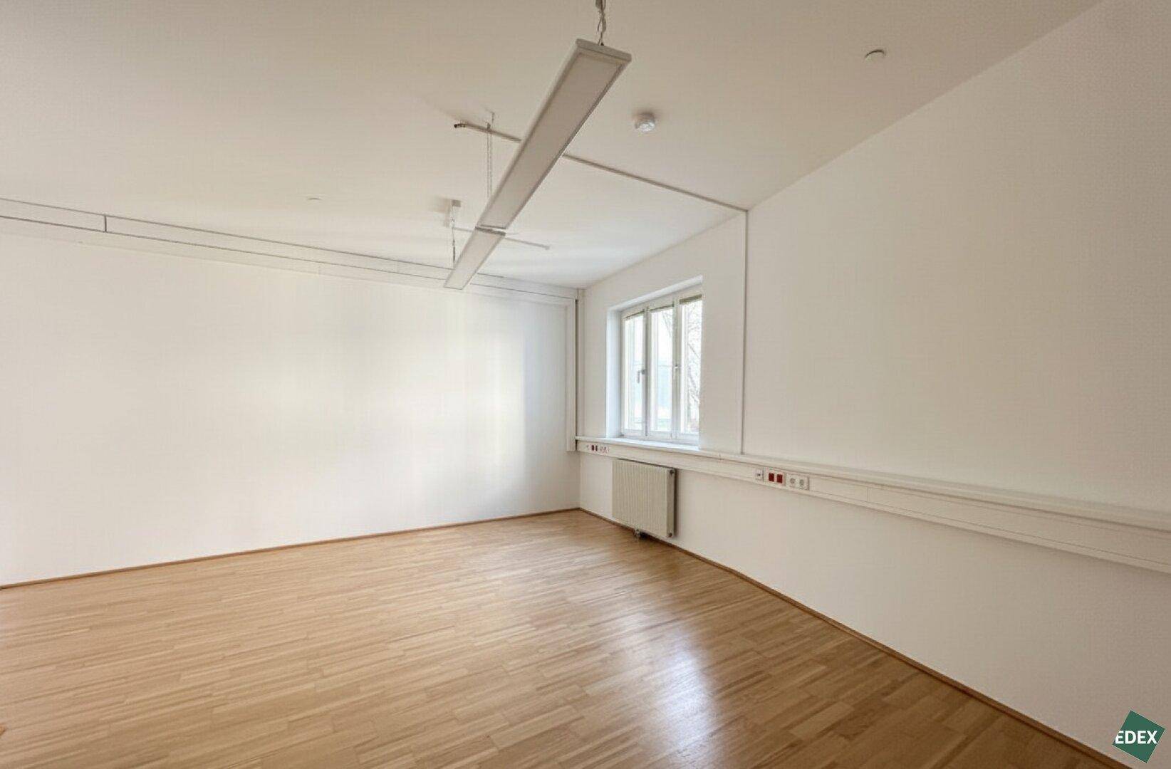 Leerer Büroraum mit einem Fenster, Heizkörper und sauberem Parkettboden, bereit zur individuellen Gestaltung.