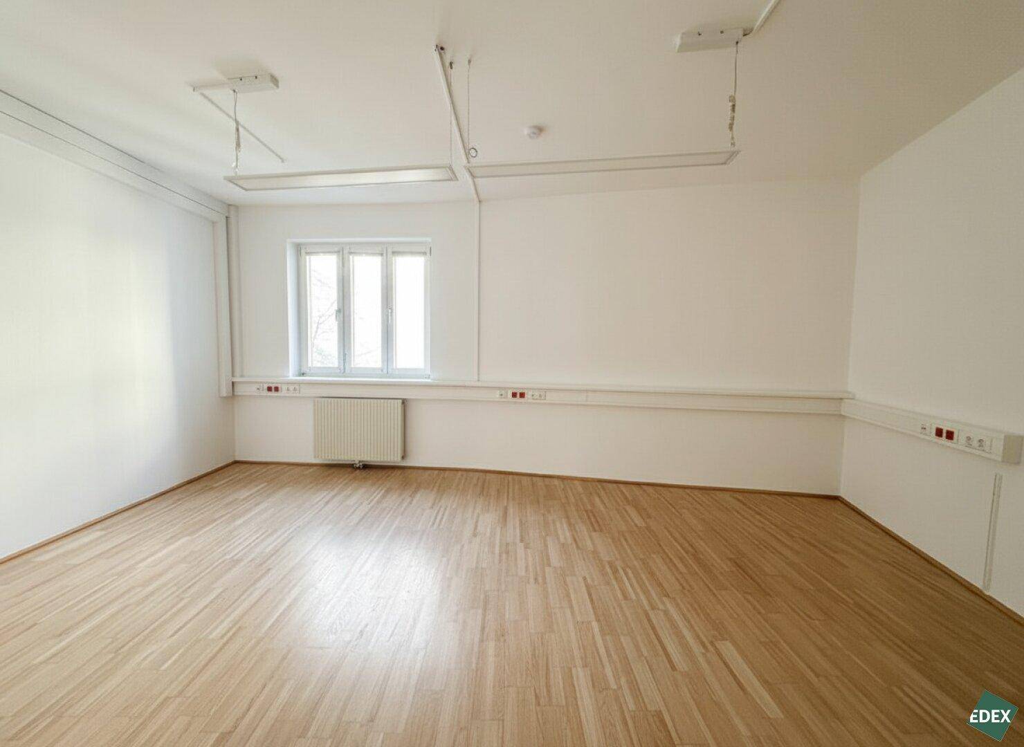 Heller Büroraum mit einem Fenster, Heizkörper und gepflegtem Parkettboden, bietet angenehme Arbeitsatmosphäre.