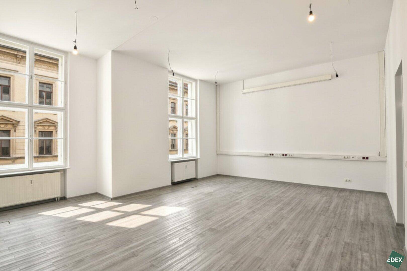 Leerer Büroraum mit hellem Holzboden, großen Fenstern und einer praktischen Wandinstallation für Stromanschlüsse.