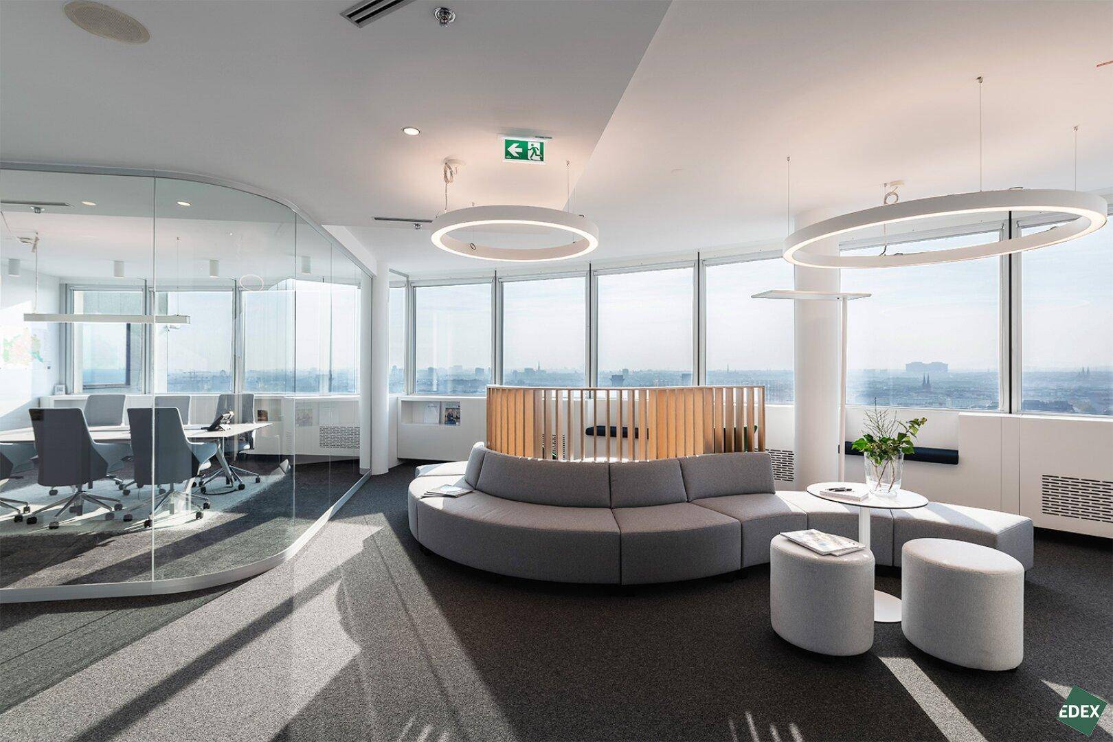 Stilvoller Büroloungebereich mit geschwungenem Sofa, Glastrennwand und weitem Blick über die Stadtlandschaft.