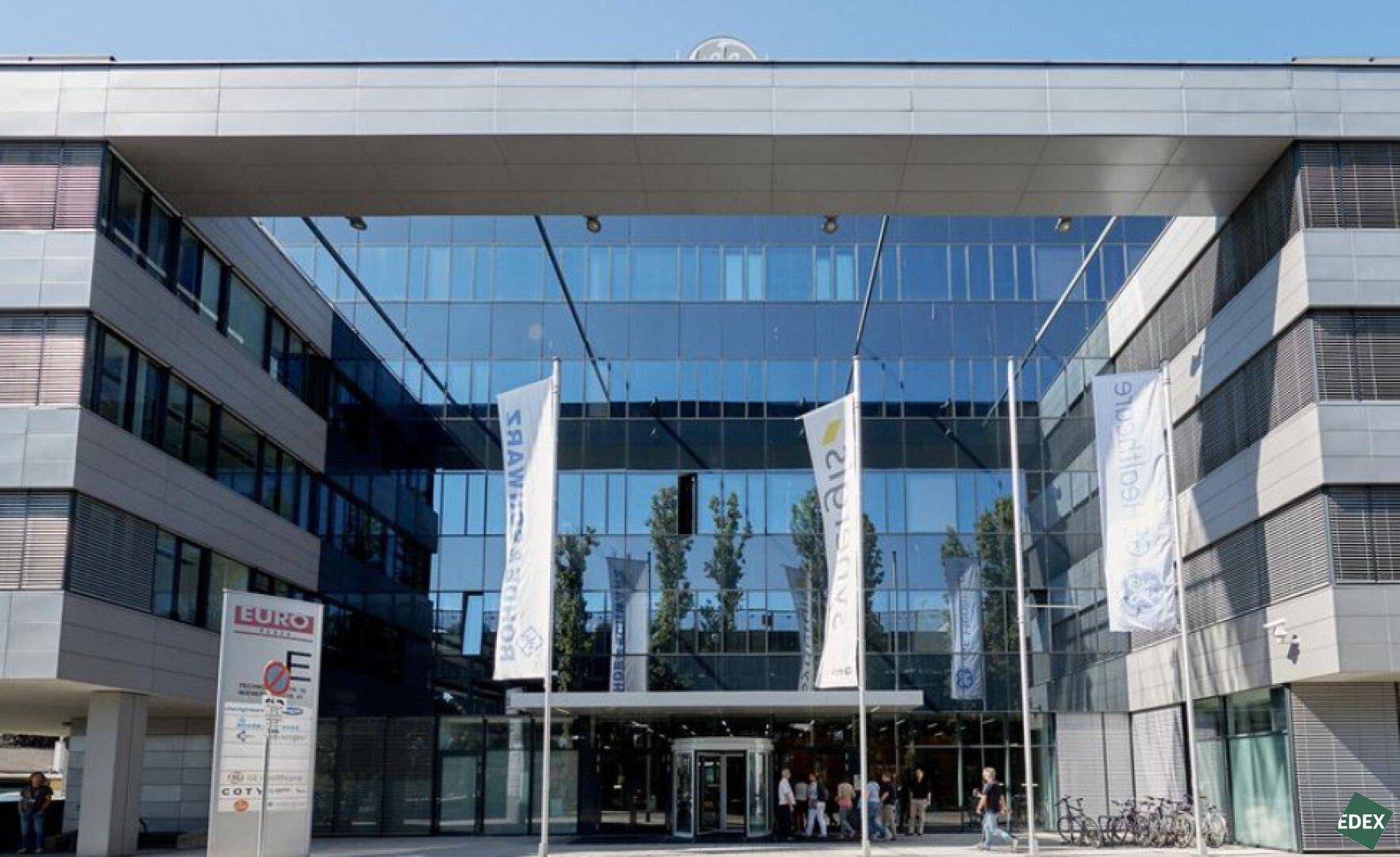 Moderne Bürogebäude-Fassade mit großer Glasfront und Eingangsbereich, umgeben von Bäumen.