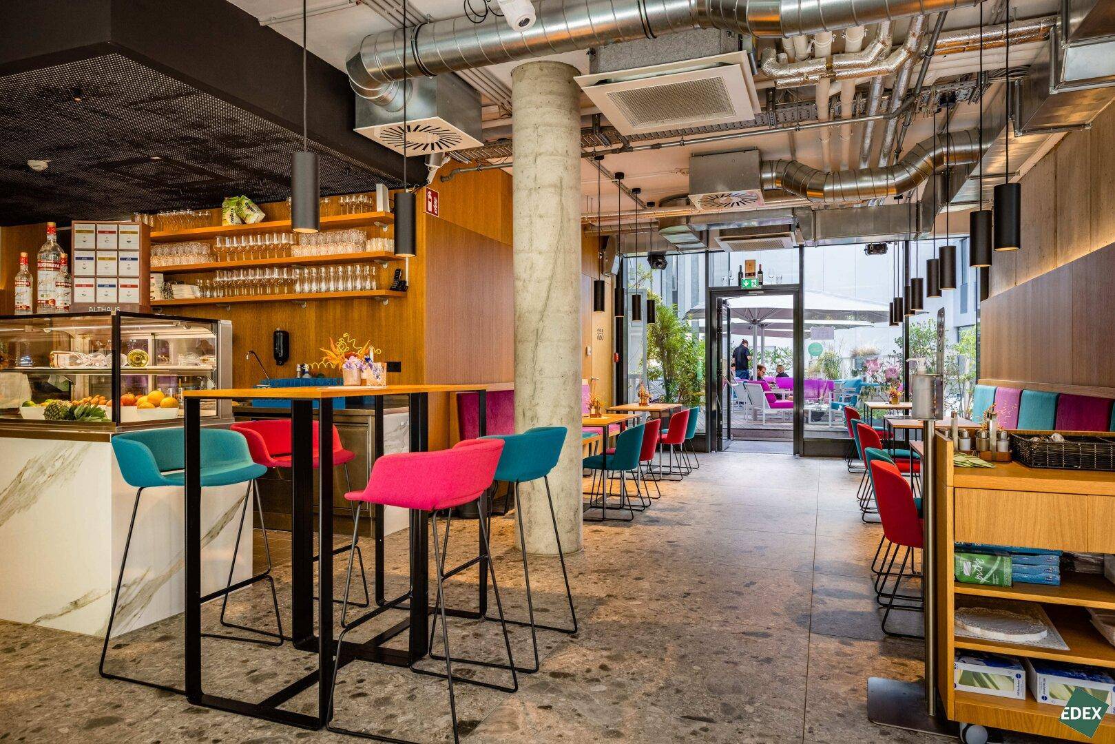 Lebhaftes Café mit bunten Stühlen, offener Decke und Zugang zu einem Außenbereich.
