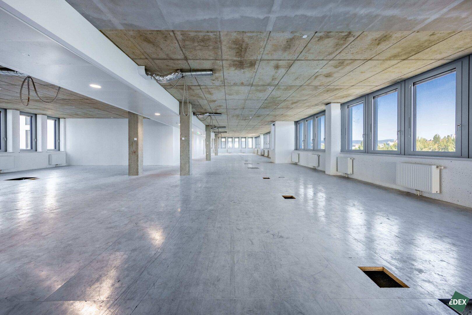 Weitläufiges Büro im Rohbau mit Betonboden, vielen Fenstern und sichtbaren Betondecken.