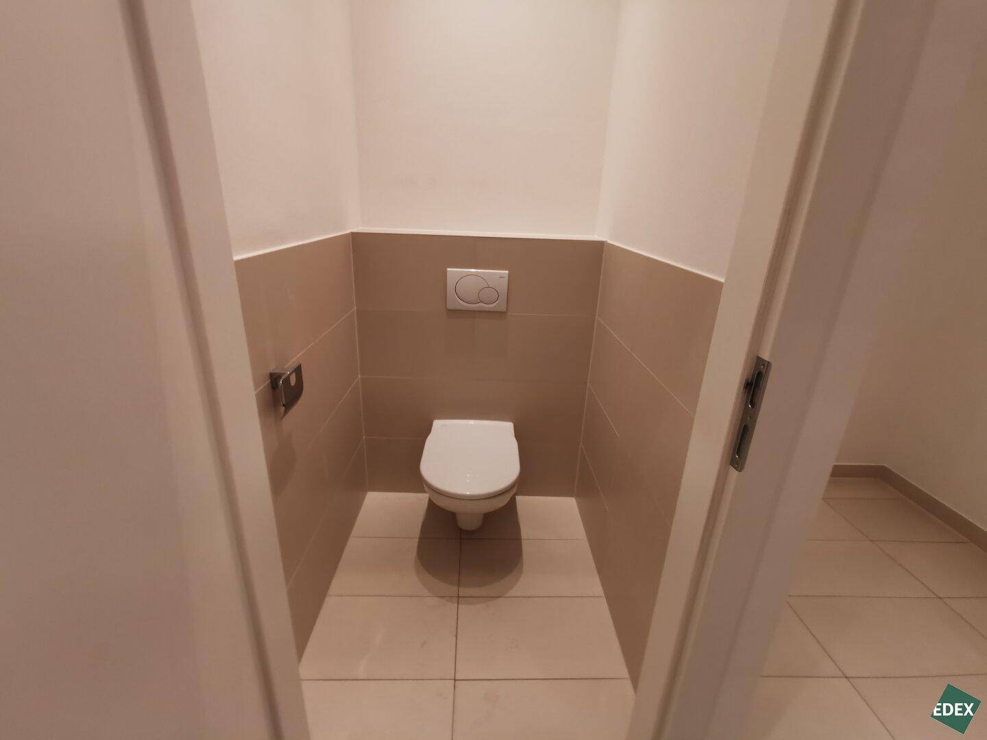 Einfache Toilette mit wandhängendem WC und beigefarbenen Fliesen, die einen sauberen Eindruck macht.