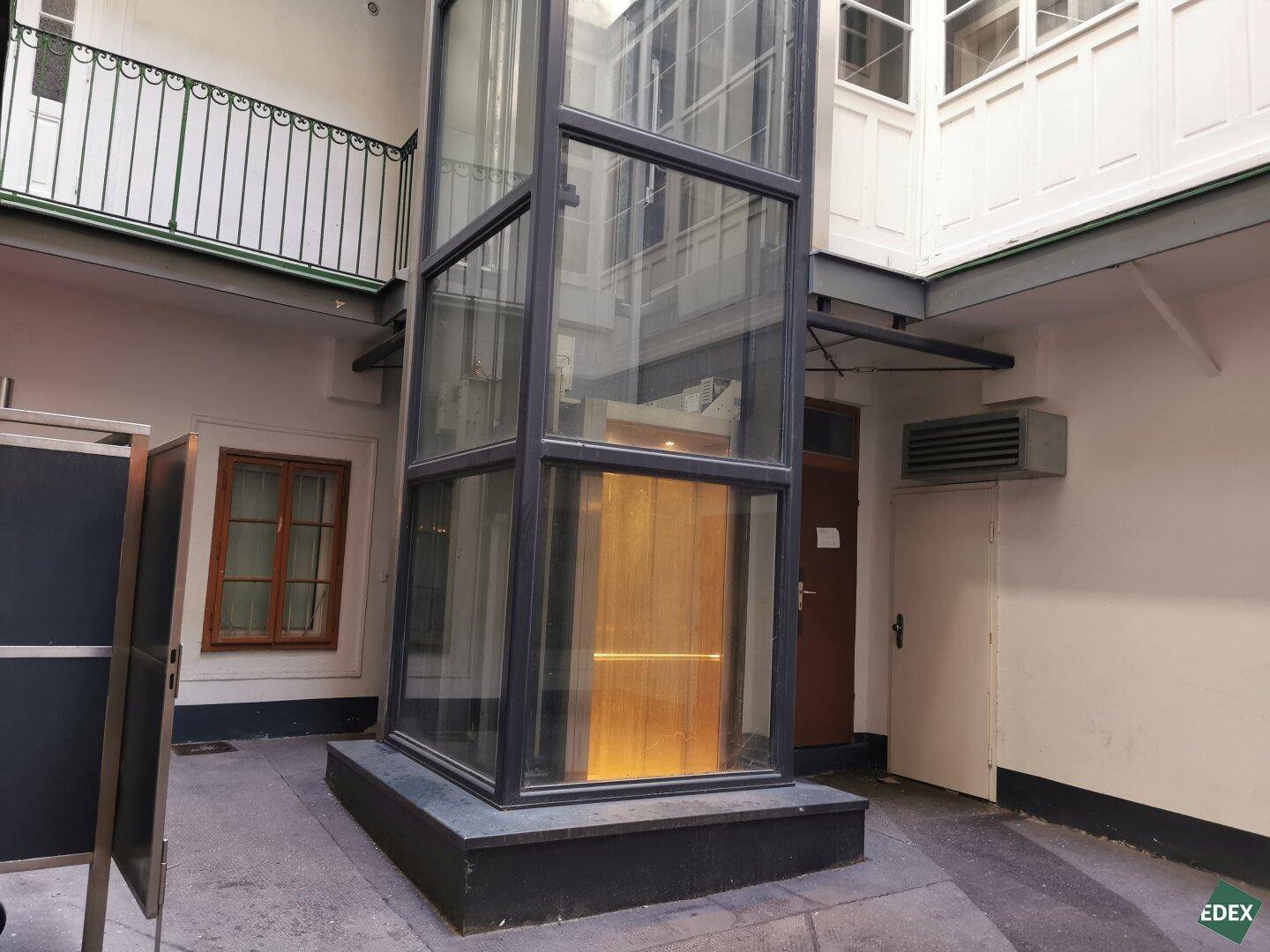 Moderner Glasaufzug im Innenhof eines Gebäudes, der eine elegante und funktionale Ergänzung darstellt.