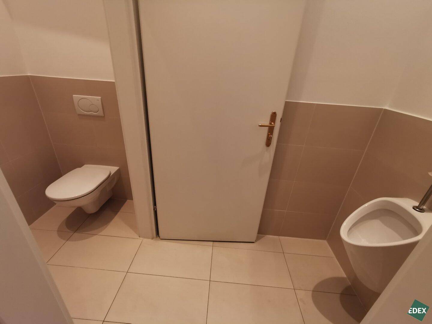 Funktionale Toilette mit wandhängendem WC und Urinal, ausgestattet mit beigefarbenen Fliesen.
