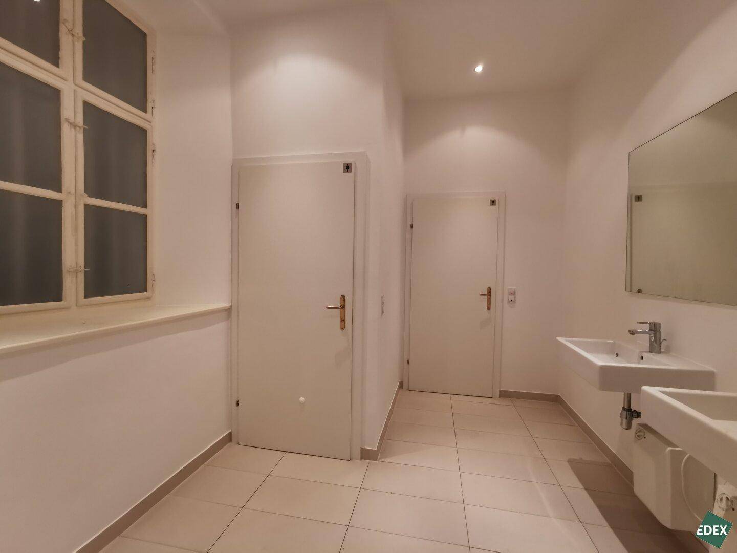 Modernes Badezimmer mit zwei Waschbecken, großem Spiegel, Fliesenboden und einem Fenster für Tageslicht.