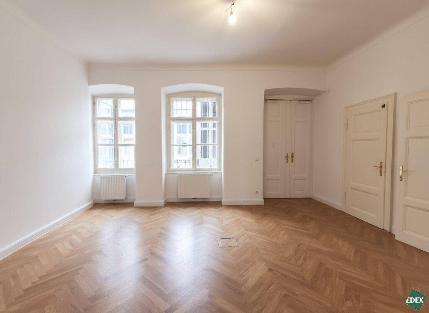 Heller Raum mit Fischgrätparkett, zwei Fenstern und zwei Türen, der vielseitig genutzt werden kann.