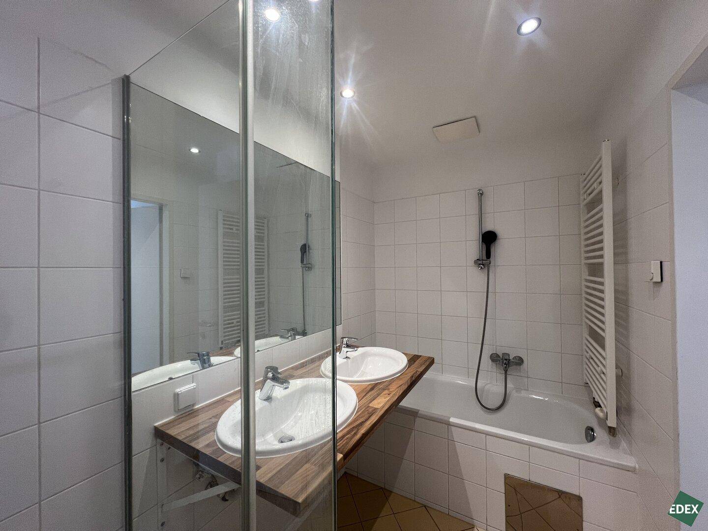 Helles Badezimmer mit Doppelwaschbecken, Badewanne und Handtuchheizkörper, gefliest in Weiß.