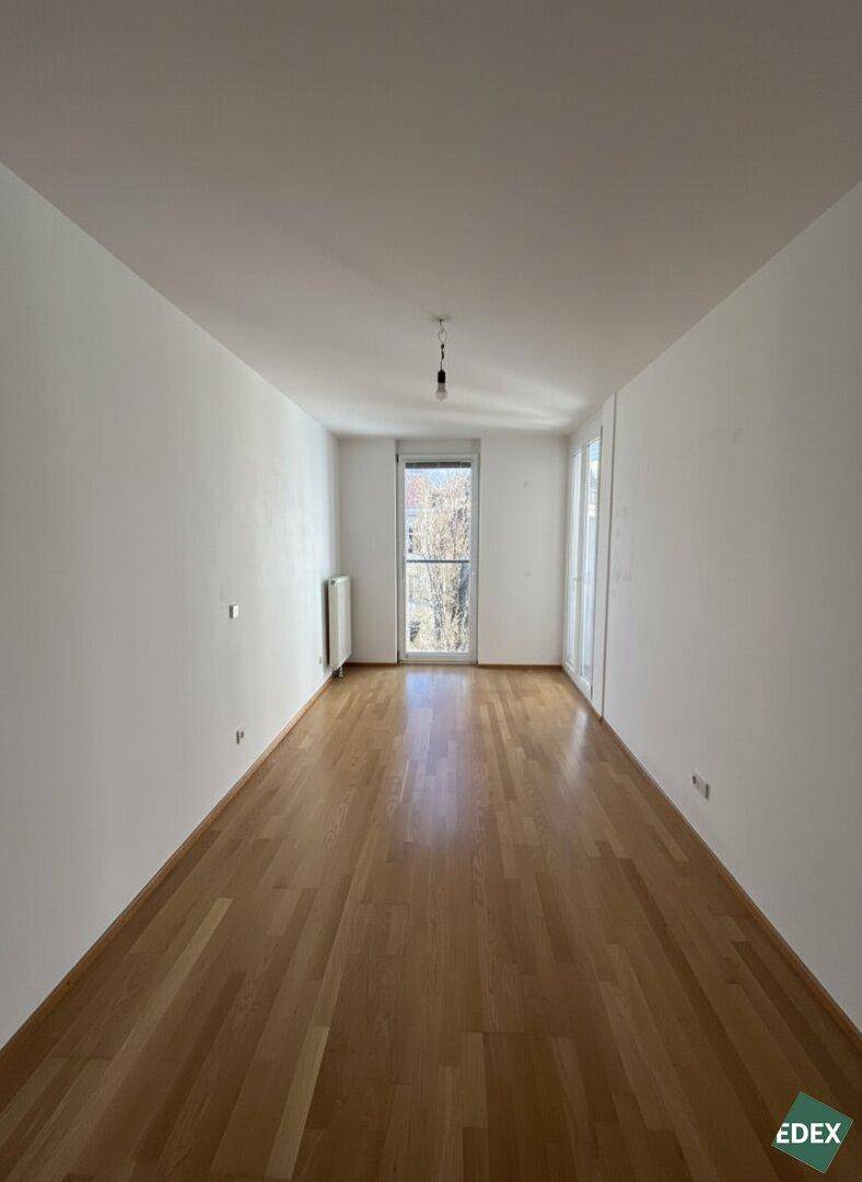 Langes, helles Zimmer mit Parkettboden und einem Fenster, das viel Tageslicht hereinlässt.