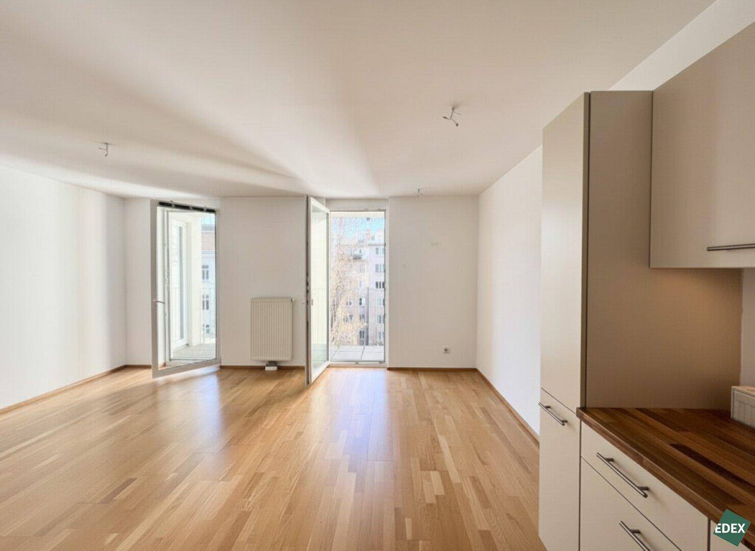 Heller Wohnbereich mit Parkettboden und Balkonzugang, angrenzend an eine kleine Küchenzeile.