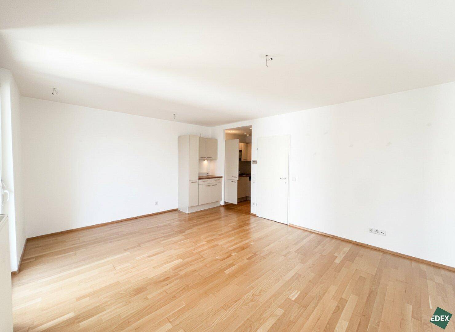 Heller Wohnraum mit Parkettboden und Zugang zur Küche, bietet viel Platz für individuelle Einrichtung.