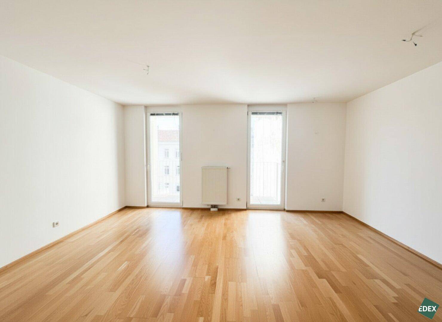 Helles Zimmer mit Parkettboden und zwei Fenstertüren, die zu einem Balkon führen.