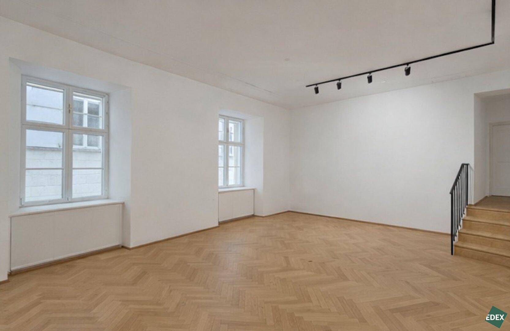 Geräumiger Raum mit Fischgrätparkett, zwei Fenstern und einer Treppe, die in eine andere Etage führt.