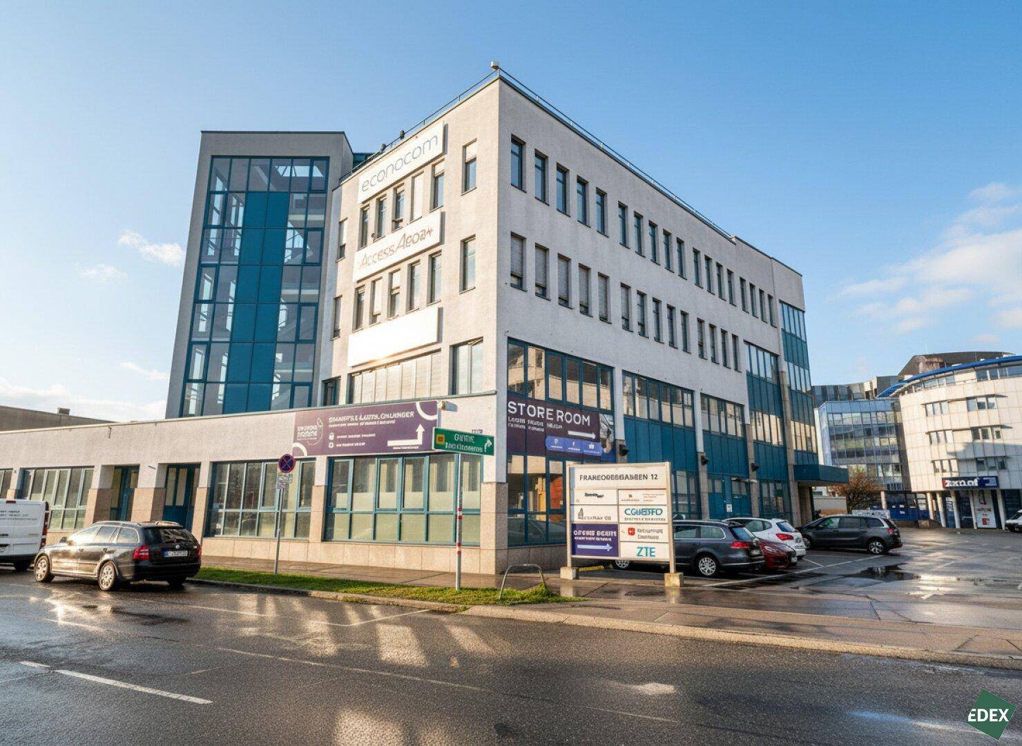 Modernes Bürogebäude mit Glasfassade und Parkplätzen im Freien. Das Gebäude beherbergt mehrere Unternehmen.