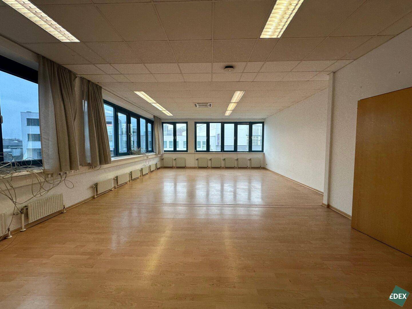 Heller, leerer Büroraum mit vielen Fenstern, die eine gute Belichtung bieten. Der Raum verfügt über einen Laminatboden und abgehängte Decken.