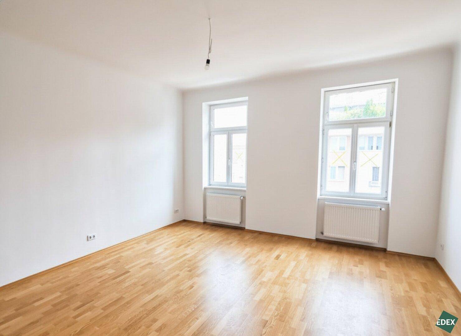 Geräumiges Zimmer mit hellem Parkettboden und großen Fenstern, ideal für Wohn- oder Schlafbereich.
